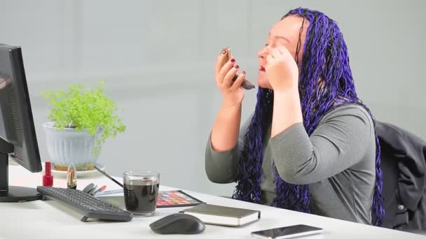 Una mujer en una oficina con un peinado afro azul se pinta los ojos con rímel mirando en un pequeño espejo al comienzo de la jornada laboral — Vídeos de Stock
