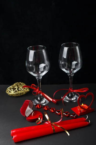 Two champagne glasses tied with ribbons on a black background next to a heart-shaped box with a ring and a candlestick with a candle and red candles. — Stock Photo, Image