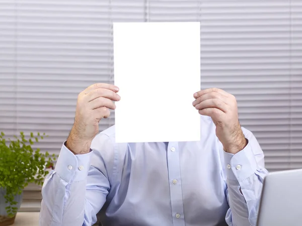 Een man in een kantoor aan een computer houdt een verticaal bord in zijn handen en bedekt zijn gezicht. — Stockfoto