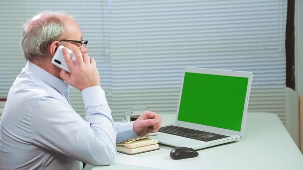 Um homem de óculos em um escritório com persianas em um computador trabalha com um projeto de tela verde e fala ao telefone. — Vídeo de Stock