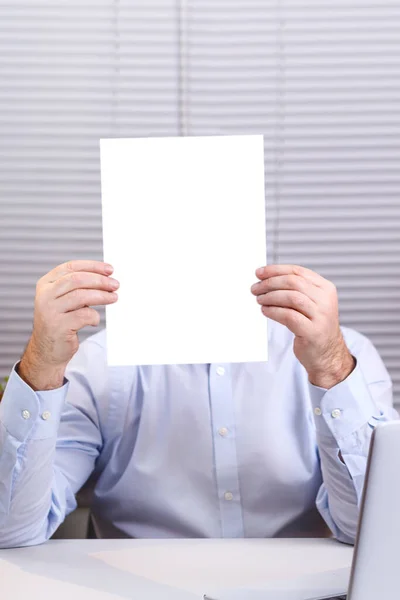 En man på ett kontor vid en dator håller en vertikal skylt i sina händer och täcker ansiktet. — Stockfoto