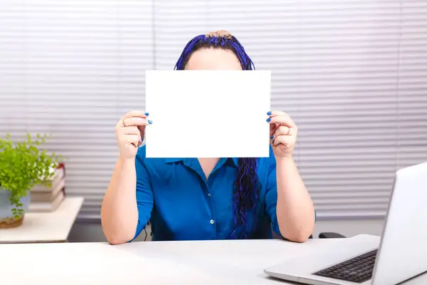 Een vrouw in het kantoor met blauwe afro vlechten aan de computer houdt een horizontale plaat in haar handen. — Stockfoto