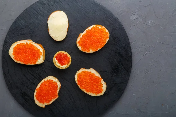 Pinceles con mantequilla y caviar rojo junto a una cuchara con caviar sobre una tabla negra sobre un fondo de hormigón. espacio de copia — Foto de Stock