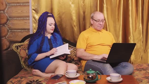 Małżeństwo w domu na kanapie przy małym stoliku mężczyzna i kobieta z afro-fryzurą czytają i bawią się w mężczyznę pracującego z laptopem — Wideo stockowe