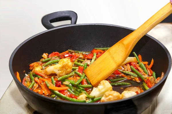 Vegetables in Pan-Asian sauce are stirred by a wooden spatula in a frying pan.