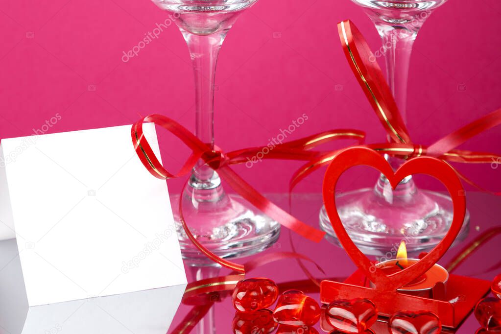 Legs of two champagne glasses with ribbons on a pink background and and a card and a heart candlestick with a burning candle.