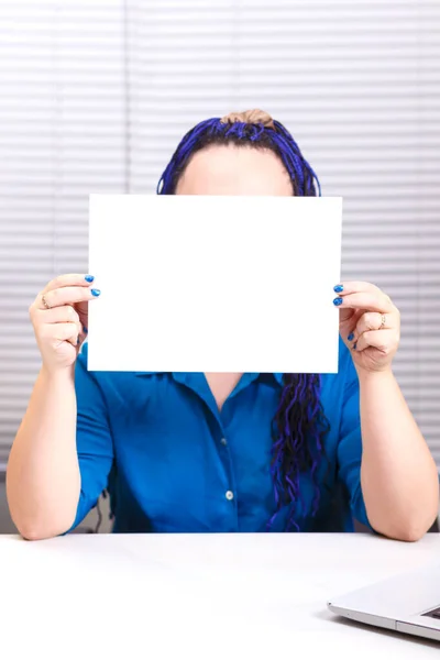 Een vrouw in het kantoor met blauwe afro vlechten aan de computer houdt een horizontale plaat in haar handen, bedekt haar gezicht. — Stockfoto