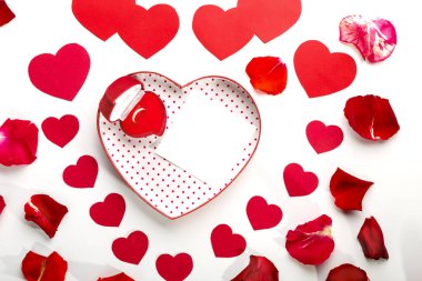 Beyaz arka planda kalp şeklinde bir kutuda altın bir yüzük. Gül yaprakları ve kırmızı kalplerin ortasında bir kart var..