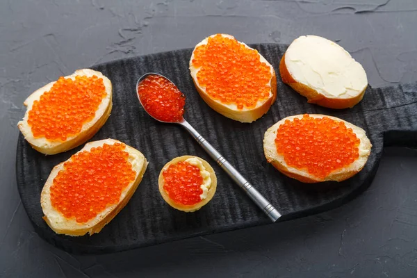 Canapés avec beurre et caviar rouge à côté d'une cuillère avec caviar sur une planche de bois sur un fond de béton. — Photo