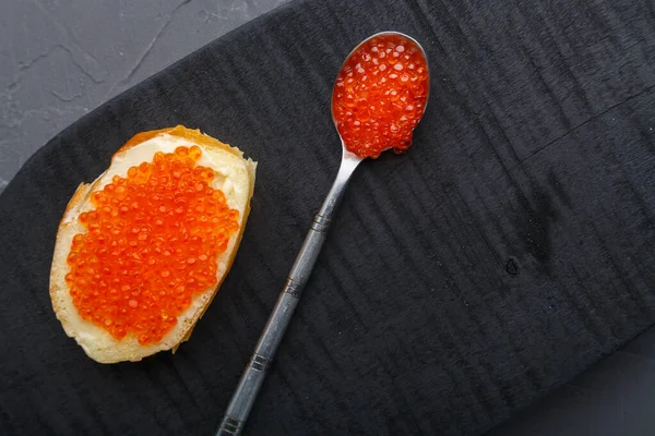 Una bruschetta con mantequilla y caviar rojo junto a una cuchara con caviar sobre una tabla de madera sobre un fondo de hormigón. — Foto de Stock