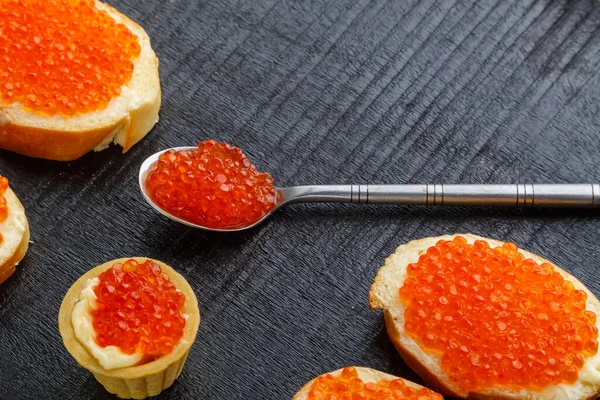 Toast con burro e caviale rosso e un cucchiaio con caviale su una lavagna nera su uno sfondo di cemento. — Foto Stock