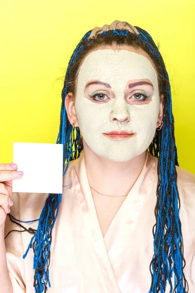 Frau mit blauen Afro-Zöpfen Gesicht in einer gefrorenen Maske aus grünem Ton mit einer Visitenkarte in den Händen auf gelbem Hintergrund. — Stockfoto