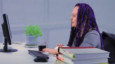 Çalışma odasında mavi afro örgülü bir kadın bilgisayar başında çalışıyor.