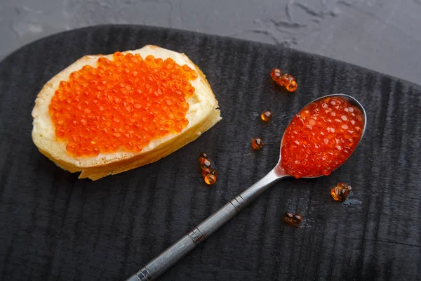 Uma torrada com manteiga e caviar vermelho ao lado de uma colher com caviar em uma tábua de madeira em um fundo de concreto. — Fotografia de Stock