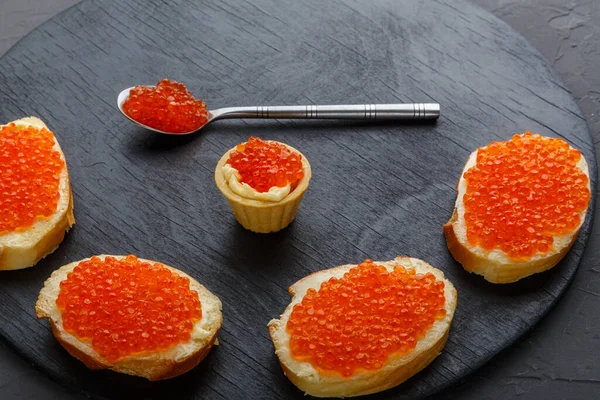 Canapés avec beurre et caviar rouge et une cuillère avec caviar sur une planche ronde noire sur un espace de copie de fond en béton — Photo