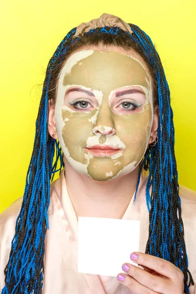 Frau mit blauen Afro-Zöpfen Gesicht in einer Maske aus grünem Ton auf gelbem Hintergrund mit einer Visitenkarte in den Händen. — Stockfoto