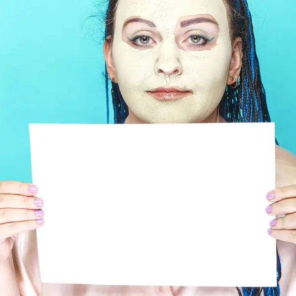 Joodse vrouw met blauwe afro vlechten gezicht in een bevroren masker van groene klei met een poster op een blauwe achtergrond. — Stockfoto