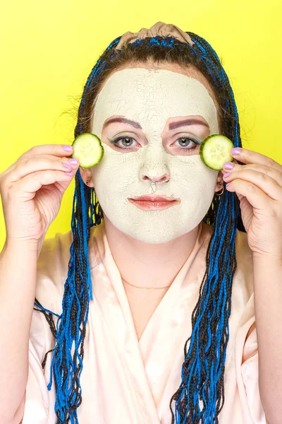 Frau mit blauen Afro-Zöpfen Gesicht in einer gefrorenen Maske aus grünem Ton mit Gurkenstücken in ihren Händen auf gelbem Hintergrund. — Stockfoto