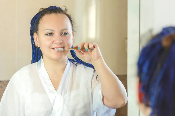 Una mujer con una túnica blanca con un peinado afro azul en el baño cerca del espejo se cepilla los dientes. —  Fotos de Stock