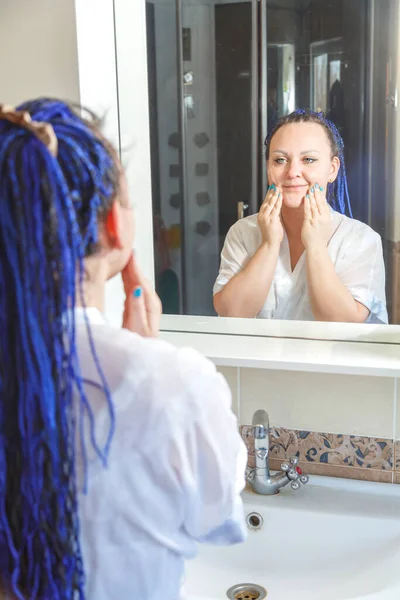 Donna in una veste bianca con un'acconciatura afro blu in bagno si riflette nello specchio fa uno scrub sul viso. — Foto Stock
