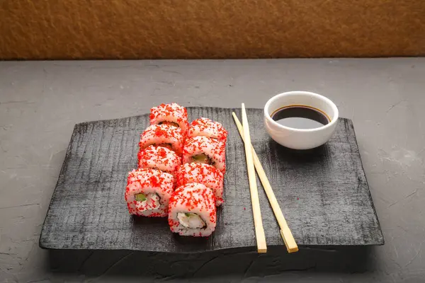 Roll with shrimp cucumber and tobiko caviar and sticks on a black board and soy sauce in a sauce bowl on a gray background. — Stock Photo, Image