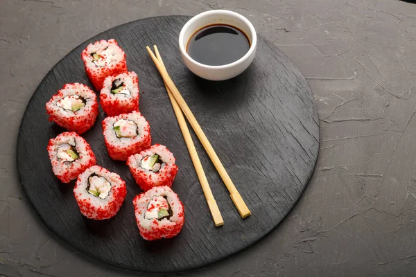 Roll s krevetami a tobiko kaviár a tyčinky na černé kulaté desce a sójovou omáčkou na šedém pozadí. — Stock fotografie