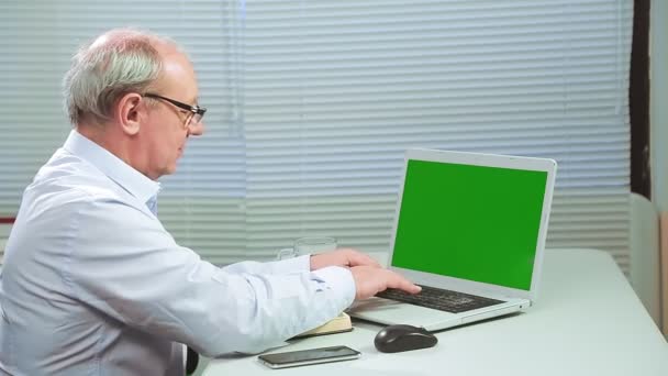 Un hombre con gafas en una oficina con persianas en una computadora trabaja una pantalla verde y habla por teléfono — Vídeos de Stock