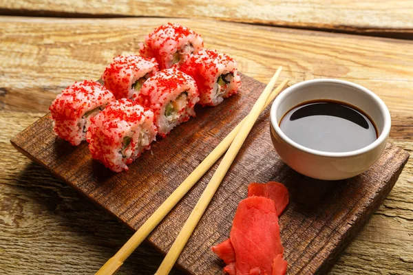 Roll with shrimp cumber and red tobiko caviar and sticks on the board and soy sauce in a sauce bowl on the wooden background. — Stock fotografie