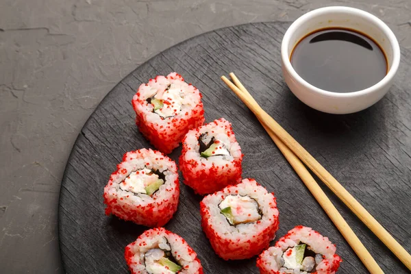 Roll with shrimp and tobiko caviar and sticks on a black round board and soy sauce on a gray background.