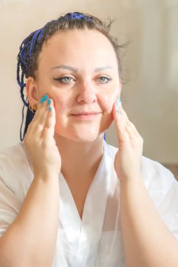 Aynanın yanındaki banyoda mavi afro saçlı beyaz cüppeli bir kadın yüz fırçası yapıyor. Dikey fotoğraf