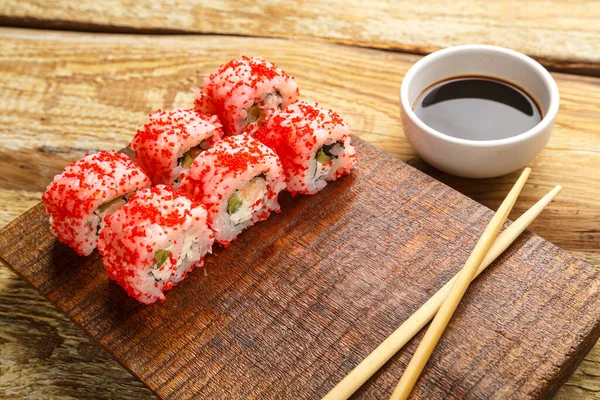 Roll with shrimp cucumber and red tobiko caviar and sticks on a board and soy sauce in a sauce bowl on a wooden background. Horizontal photo