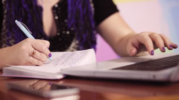 Wanita muda dengan Afrokos biru di rumah di sofa di meja dengan laptop menulis informasi dalam sebuah buku catatan tanpa wajah — Stok Video