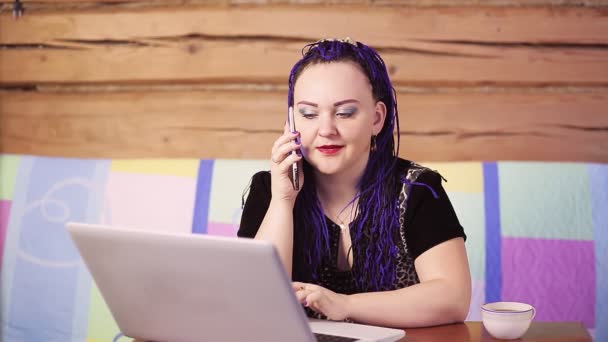 Jonge vrouw met blauwe Afrokos thuis op de bank aan tafel met een laptop spreekt aan de telefoon. — Stockvideo