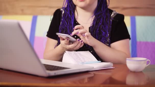 A young woman with blue Afrokos at home on the couch at a table with a laptop is texting in a smartphone in instant messengers — Stock Video