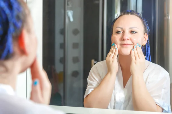 Aynanın önündeki banyoda mavi afro saçlı beyaz cüppeli bir kadın yüzünü ovuyor..