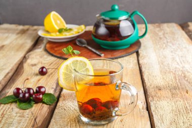 Naneli bir fincanda siyah çay ve taze kirazların yanında ahşap bir masada limon ve tezgahta bir çaydanlık ve tabakta limon..