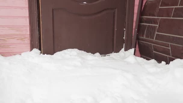 Uma mulher com um casaco de inverno com dificuldade abre a porta da casa após uma queda de neve, limpando a neve com o pé e deixando a casa — Vídeo de Stock