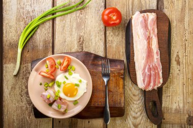 Çırpılmış yumurta, domuz pastırması, domates ve soğanlı ve ahşap bir masada, çatal, dilimlenmiş pastırma ve domatesin yanında..
