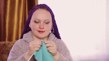 Mavi afro saçlı Yahudi bir kadın odadaki kanepeye örgü örüyor.