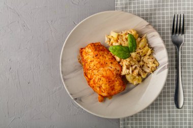 Fırınlanmış tavuk göğsü gri bir tabakta, ananas salatası masanın üstünde, peçetenin üzerinde, beton bir arka planda, çatalın yanında..