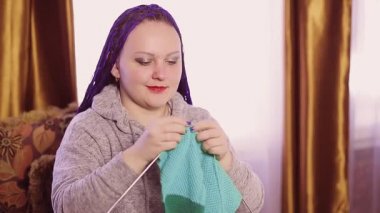 Mavi afro saçlı Yahudi bir kadın odadaki kanepeye örgü örüyor.