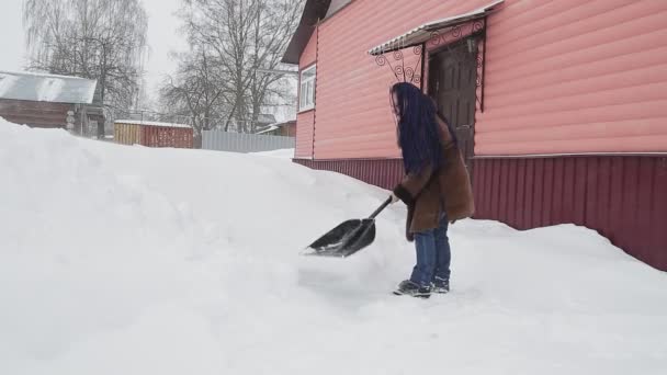 Una donna in un cappotto caldo con trecce afro sciolto con una pala rimuove la neve dopo una nevicata nel cortile della sua casa — Video Stock