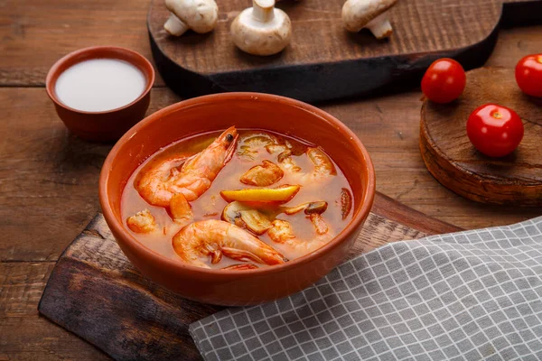 Tom yam soup with shrimps in a plate on the table on a napkin next to a bowl of coconut milk mushrooms and tomatoes.