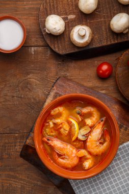 Tom yer elması çorbası, karidesli ve ahşap bir masada, peçetenin üzerinde hindistan cevizi sütü mantarlı ve domatesli bir kasenin yanında..