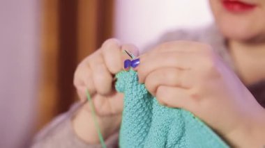 Yüzü olmadan fotoğraf çeken bir kadın elinde örgü iğneleriyle basit bir desenle süveter örüyor.