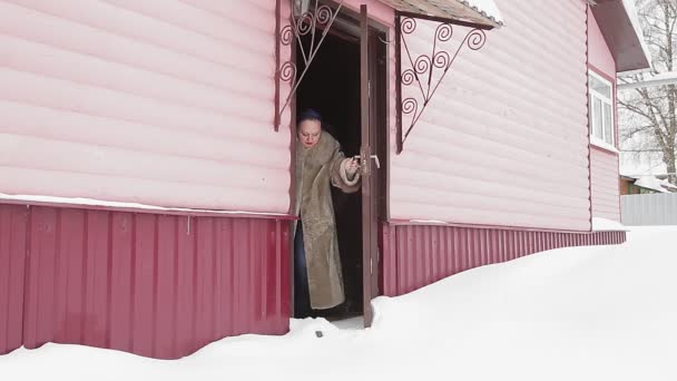 Une femme dans un manteau d'hiver avec difficulté ouvre la porte de la maison après une chute de neige, pelletant la neige avec son pied et sort dans la cour — Video