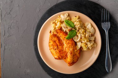 Bej bir tabakta fırında tavuk göğsü, yuvarlak bir masada ananas salatası, beton bir arka planda, çatalın yanında..