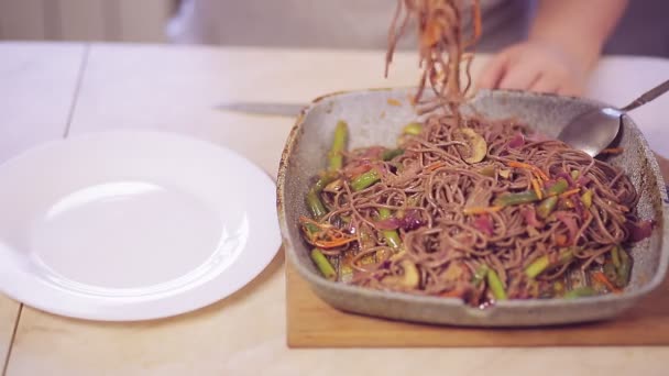 A womans hand with tongs transfers soba with mushrooms and vegetables from a frying pan to a plate. — Stock Video