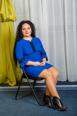 Mavili kıvırcık esmer kadın bacaklarını uzatmış bir sandalyede oturuyor..