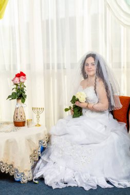 Yüzü bir buket beyaz gülle örtülü mutlu bir Yahudi gelin Huppa ayinini çiçeklerle dolu bir masada yapmadan önce sinagogda oturur..